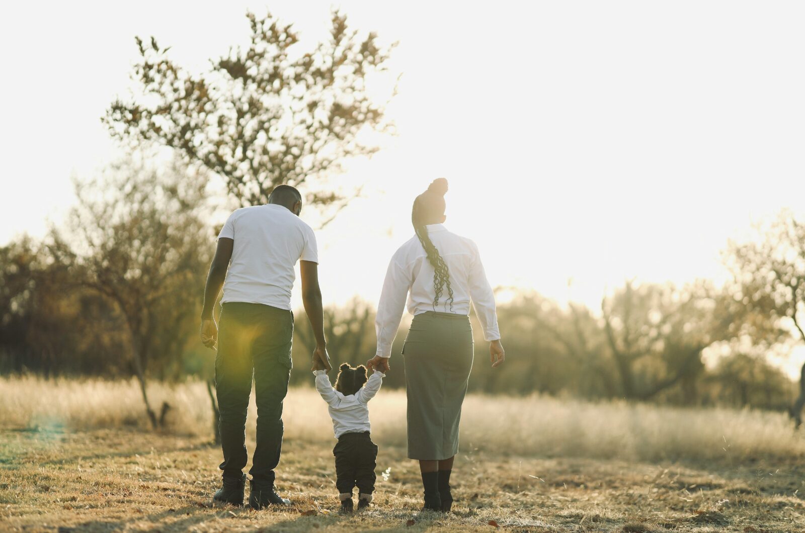 Two adults walking hand in hand with a small child through a sunlit grassy field, enjoying a peaceful outdoor moment.