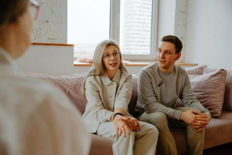 Couple sitting on a couch talking with a therapist, during a counseling session in a calm, well-lit office setting.
