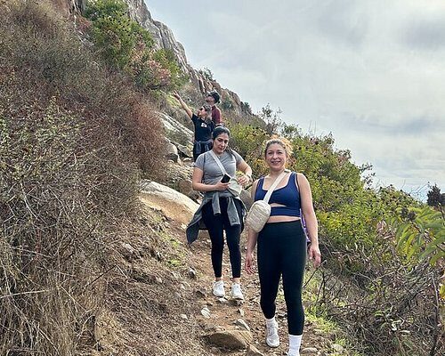 Group of hikers walking along a narrow mountain trail surrounded by rocks and greenery under a cloudy sky.